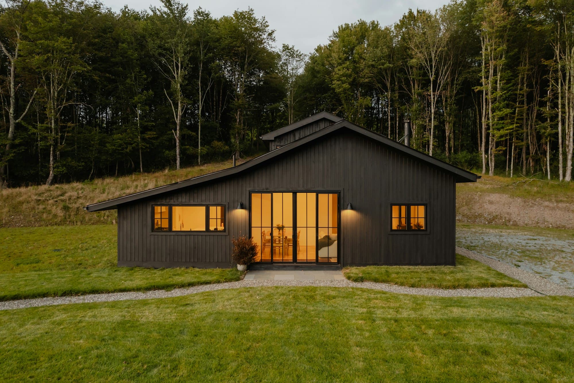 A dark timber-clad garden building at dusk, glazed doors lit warm against the lawn and trees