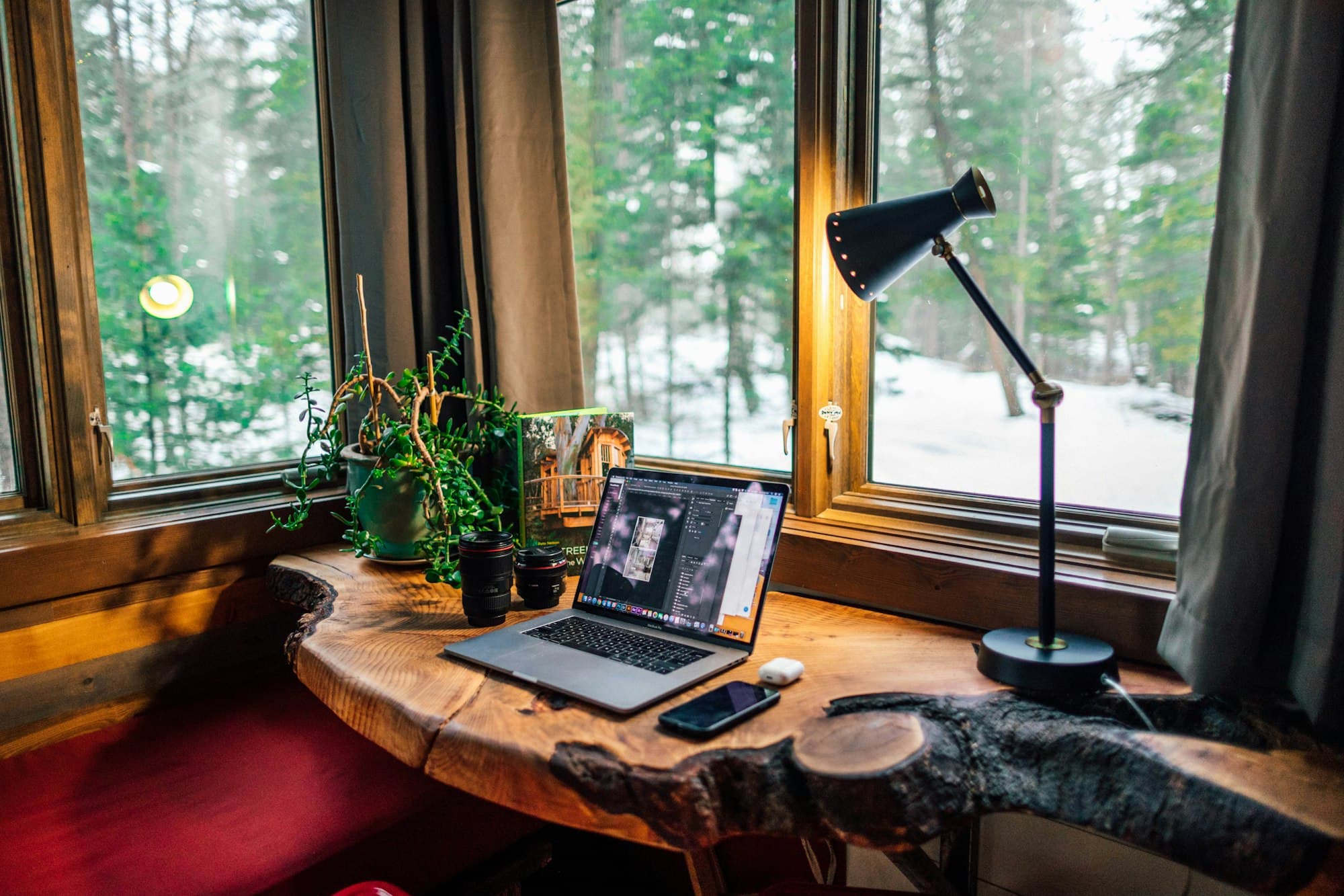 A laptop on a timber desk beneath a wide window looking onto trees